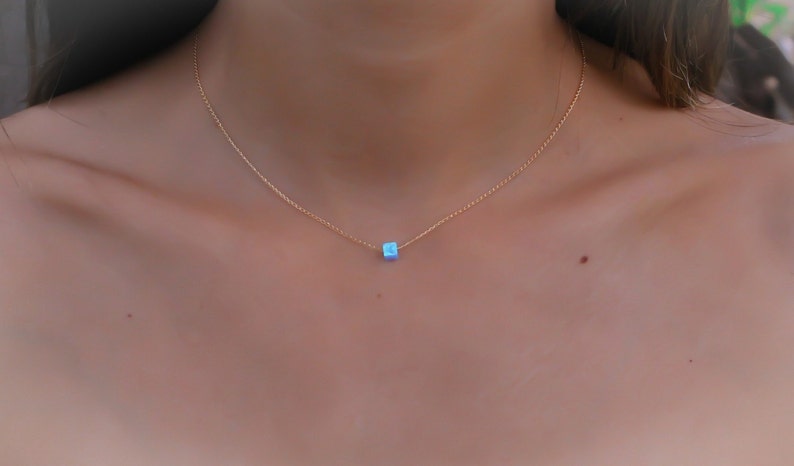 May include: A delicate gold necklace featuring a small, square, light blue pendant. The pendant is centered on a thin gold chain, creating a minimalist and elegant design. The necklace is shown against a skin tone background.