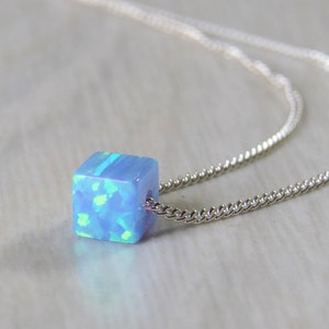 May include: A silver necklace featuring a small, light blue cube pendant with an iridescent, opal-like appearance. The cube is suspended from a delicate silver chain, creating a minimalist and elegant design. The pendant has a subtle, textured surface.