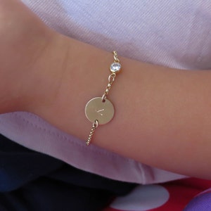 Könnte beinhalten: Ein goldenes Kettenarmband mit einem kleinen runden, klaren Stein-Anhänger und einem größeren runden Anhänger mit dem eingravierten Buchstaben "L".