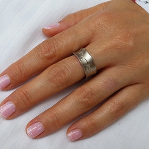 Puede incluir: Un anillo de plata con un dise&ntilde;o ondulado texturizado. El anillo se lleva en el dedo anular de una mano con esmalte de u&ntilde;as rosa.