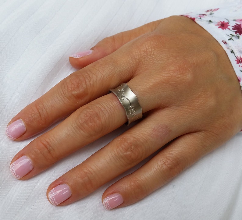 Puede incluir: Un anillo de plata con un dise&ntilde;o ondulado texturizado. El anillo se lleva en el dedo anular de una mano con esmalte de u&ntilde;as rosa.