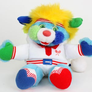May include: A plush toy in the shape of a cartoon character with a blue face, yellow hair, and green eyes. It wears a white outfit with red and blue accents, and a red headband. The toy has a red nose and a white ball.