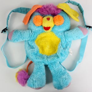 May include: A plush, blue backpack shaped like a cartoon animal with a yellow belly, orange ears, and a purple and yellow tuft of hair. The backpack has blue straps and a colourful tail. The plush toy has a red nose and pink accents.
