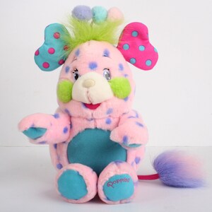 Könnte beinhalten: Ein rosa Plüschtier mit blauen und rosa Punkten, grünen flauschigen Ohren und einem blau-violetten Schwanz. Das Spielzeug hat ein weißes Gesicht mit blauen Augen und einem rosa Mund. Das Spielzeug sitzt auf einer weißen Oberfläche und hat das Wort "Goppit!" auf seinem Fuß.