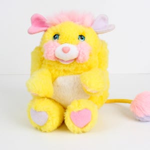 May include: A plush yellow toy animal with pink and purple accents. The toy has a fluffy yellow body, pink ears, and a pink and purple tail. The toy's feet have heart-shaped details. The toy has blue eyes and a pink nose.