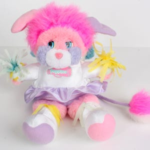 May include: A colorful plush toy with pink, purple, and blue accents. The toy has a fluffy pink head, long ears, and a white dress with a purple skirt. It holds pom-poms and has a fluffy tail. The word "Popples" is on the dress.