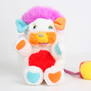 May include: A colorful plush toy with a white body, a pink mohawk, and accents of orange, blue, and yellow. The toy has a red heart-shaped patch on its chest and a yellow pom-pom tail. It is a vintage collectible.
