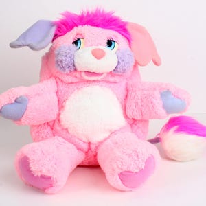 May include: A plush pink stuffed animal with a dog-like appearance. It has a white belly, blue eyes, and accents of purple and pink. The toy has a fluffy tail and is designed to be a backpack.