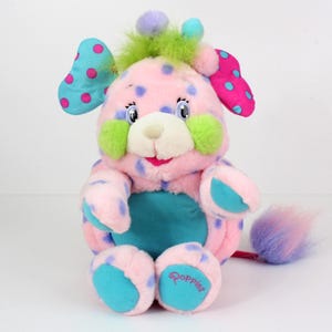 May include: A pink and blue plush toy with a green mohawk and a blue tail. The toy has a white face with blue eyes and a pink nose. The toy is sitting on a white surface. The toy is labeled "Popples".