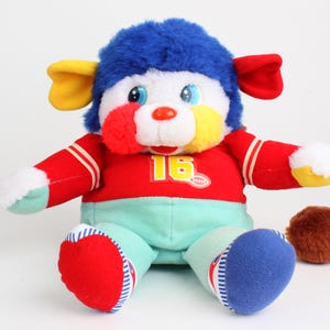 May include: A colorful plush toy with a blue furry head, yellow and red ears, and a red and white striped shirt. The toy has a white face with blue eyes, a red nose, and a yellow number 16 on the shirt. The toy is sitting with a brown ball next to it.