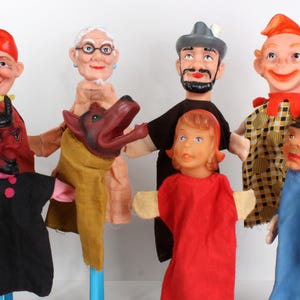 May include: A collection of vintage hand puppets featuring various characters. The puppets have painted faces and fabric clothing. Characters include a devil, an elderly woman, a dog, a man with a beard, a young girl, a jester, and a man in a blue uniform.