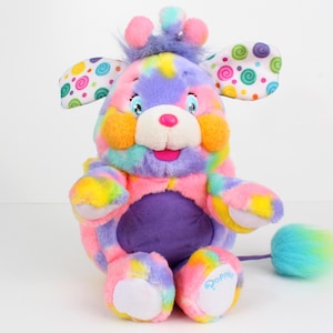 May include: A colourful plush giraffe toy with a rainbow tie-dye pattern. The toy has large ears with polka dots, orange cheeks, a pink nose, and a blue and turquoise tail. The word "Poppy" is written on one foot.