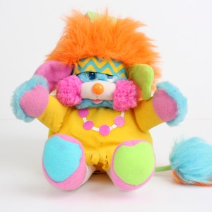 May include: A brightly coloured plush toy with a yellow body, pink and blue details, and an orange mane. The toy has a blue face with a zigzag pattern, pink fur around the cheeks, and a pink and white necklace. The tail is blue and orange.
