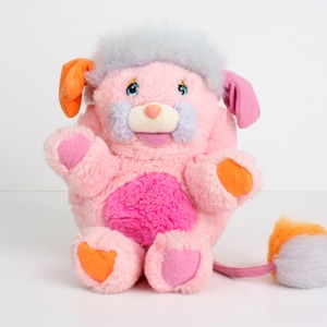 May include: A plush pink dog-like stuffed animal with a light blue head of fur, orange ears, and a pink tail. The toy has a pink body with a darker pink heart-shaped patch on its belly, and orange and pink accents on its paws.