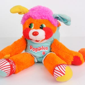 May include: A colorful Popples plush toy. The plush has orange fur, a pink head, and blue ears. The toy has a red nose, green eyes, and red cheeks. The plush is wearing a light blue shirt with the word "Popples" on it. The feet are striped.