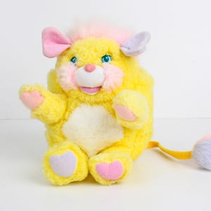 May include: A vintage yellow plush toy with pink and purple accents. The toy has a white belly, blue eyes, and a pink nose and cheeks. It has a yellow tail with a fluffy, multi-colored tip. The toy is in a sitting position with one arm raised.
