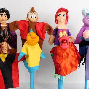 Puede incluir: Una colecci&oacute;n de marionetas de personajes coloridos, incluyendo a Aladd&iacute;n, un mono, Flounder, Ariel, &Uacute;rsula y un caballo. Cada marioneta tiene una cara detallada y viste ropa vibrante. Las marionetas est&aacute;n montadas en palos azules.