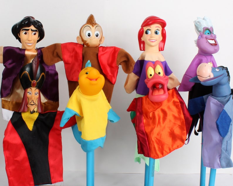 Puede incluir: Una colecci&oacute;n de marionetas de personajes coloridos, incluyendo a Aladd&iacute;n, un mono, Flounder, Ariel, &Uacute;rsula y un caballo. Cada marioneta tiene una cara detallada y viste ropa vibrante. Las marionetas est&aacute;n montadas en palos azules.
