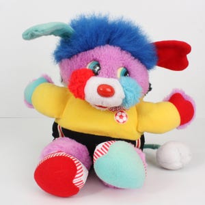 May include: A plush toy in the shape of a dog wearing a yellow and black football kit. The toy has blue hair, pink fur, and a red nose. It is holding a white football.