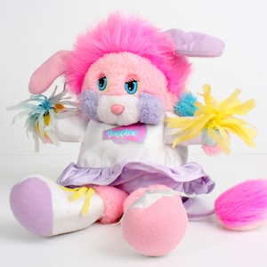 May include: A plush pink and white bunny doll with a pink mane, blue eyes, and a pink nose. The doll wears a white dress with a lavender skirt and holds pom-poms. The doll has the word "Popples" on the front.