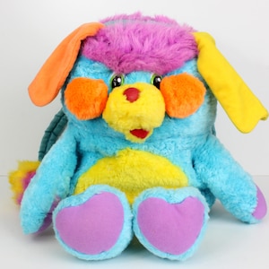 Peut inclure: Une peluche colorée en forme d'animal de dessin animé. Elle présente un corps bleu, un ventre jaune et des pieds violets. Le jouet a des joues oranges, un nez jaune et une tête rose et violette. Les oreilles sont orange et jaune.