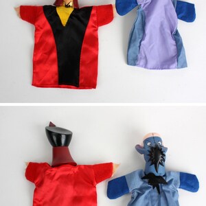 Puede incluir: Dos marionetas de mano con personajes de dibujos animados. Una marioneta tiene un atuendo rojo y negro con un sombrero negro. La otra marioneta es azul y morada. Las marionetas est&aacute;n sobre un fondo blanco.