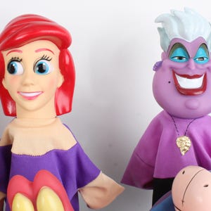 Puede incluir: Dos mu&ntilde;ecas de personajes de la pel&iacute;cula de animaci&oacute;n, La Sirenita. Ariel tiene el pelo rojo y una camisa morada y beige. &Uacute;rsula, la bruja del mar, es morada con pelo blanco y un collar dorado. Las mu&ntilde;ecas est&aacute;n sobre un fondo blanco.