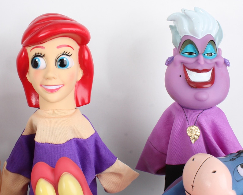 Puede incluir: Dos mu&ntilde;ecas de personajes de la pel&iacute;cula de animaci&oacute;n, La Sirenita. Ariel tiene el pelo rojo y una camisa morada y beige. &Uacute;rsula, la bruja del mar, es morada con pelo blanco y un collar dorado. Las mu&ntilde;ecas est&aacute;n sobre un fondo blanco.