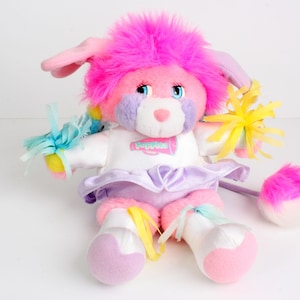 May include: A soft toy in pastel shades, with a pink body, purple details, and a fluffy pink mane. The toy has a white top with the word "Poppies" and a lavender skirt. It has a tail and is decorated with ribbons.