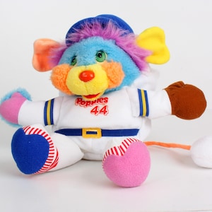 May include: A colourful plush toy in a baseball uniform. The toy has a blue hat, multi-coloured fur, and a yellow nose. The white uniform has the word "Poppies" and the number "44" on the front. The toy has a pink and blue baseball and a white ball.