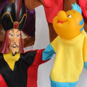 Puede incluir: Primer plano de dos marionetas de personajes de Disney. Una marioneta es Jafar, con un sombrero negro, t&uacute;nicas rojas y negras y una expresi&oacute;n severa. La otra es una marioneta de pez amarillo con detalles azules, que lleva una camisa amarilla.