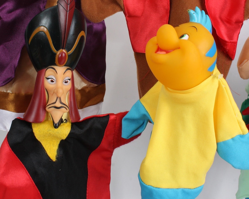 Puede incluir: Primer plano de dos marionetas de personajes de Disney. Una marioneta es Jafar, con un sombrero negro, t&uacute;nicas rojas y negras y una expresi&oacute;n severa. La otra es una marioneta de pez amarillo con detalles azules, que lleva una camisa amarilla.