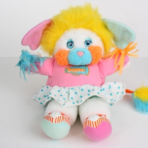 May include: A vintage soft toy with a white body, pink dress, and yellow hair. The toy has blue eyes, orange cheeks, and a blue nose. The dress has a white skirt with blue polka dots. The toy's feet are teal and pink.