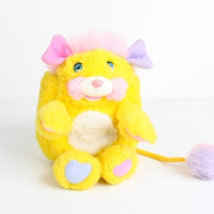 May include: A vintage yellow plush toy with pink and purple accents. The toy has pink ears, blue eyes, and a pink nose. It features a fluffy pink and purple tail and a white belly. The toy is a retro collectible.