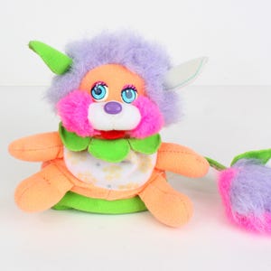 May include: A brightly coloured plush toy with a flower-shaped tail. The toy has a lavender head, orange body, and pink accents. It features blue eyes, a green collar, and a white and yellow patterned belly. The tail is purple and pink.