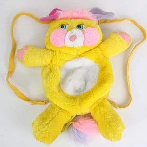 May include: A plush yellow backpack shaped like a cartoon animal with pink cheeks, ears, and paw pads. The backpack has a white belly, blue eyes, and a multi-colored tail. It features yellow straps for carrying.