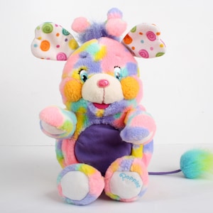 May include: A colourful plush toy with a purple, pink, yellow and blue patchwork design. The toy has large, floppy ears with a white background and colourful polka dots. The toy has a purple belly and a long, fluffy tail with green and blue tips. The toy is labelled "Popples" on the bottom.