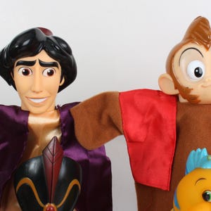Puede incluir: Dos mu&ntilde;ecos de personajes de la pel&iacute;cula de animaci&oacute;n Aladdin. Uno es Aladdin, con un chaleco morado y una camisa dorada. El otro es un mono con un chaleco rojo. Tambi&eacute;n se ve un mu&ntilde;eco de pez amarillo.