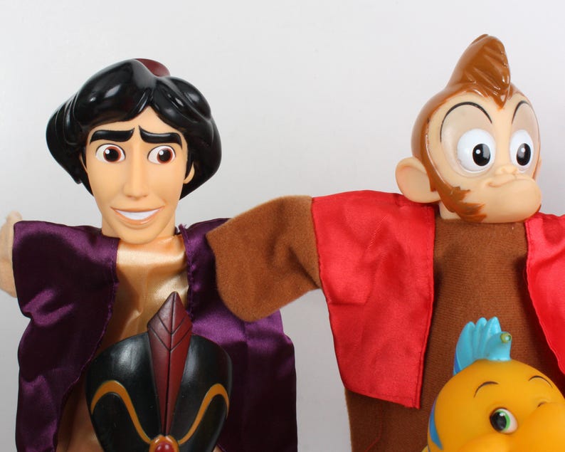 Puede incluir: Dos mu&ntilde;ecos de personajes de la pel&iacute;cula de animaci&oacute;n Aladdin. Uno es Aladdin, con un chaleco morado y una camisa dorada. El otro es un mono con un chaleco rojo. Tambi&eacute;n se ve un mu&ntilde;eco de pez amarillo.
