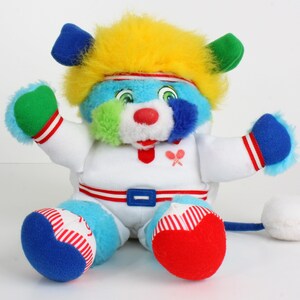 May include: A vibrant plush bear toy with blue, yellow, green, and red elements. The bear is dressed in a white outfit with red and blue details, a red belt, and a blue tail. It has a red nose and a tennis racket emblem.