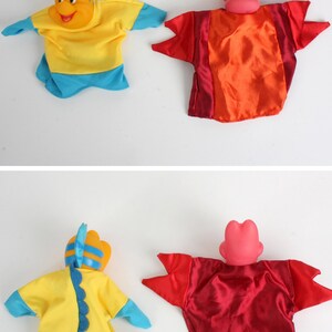 Puede incluir: Dos marionetas de mano de tela: un personaje amarillo y azul con forma de pez y un personaje rojo y naranja con forma de cangrejo. Las marionetas tienen cuerpos de tela y cabezas de pl&aacute;stico, probablemente para el juego infantil.