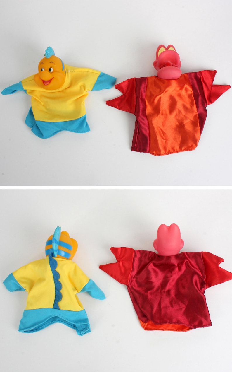 Puede incluir: Dos marionetas de mano de tela: un personaje amarillo y azul con forma de pez y un personaje rojo y naranja con forma de cangrejo. Las marionetas tienen cuerpos de tela y cabezas de pl&aacute;stico, probablemente para el juego infantil.