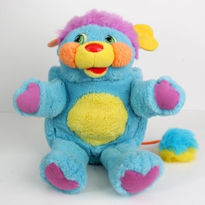 May include: A vintage plush toy in the shape of a friendly creature. It has a light blue body, a yellow belly, and purple accents on its head, paws, and feet. The toy features orange cheeks, a yellow nose, and a red mouth. A small, fluffy ball is attached.