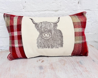 May include: Rectangular decorative pillow featuring a Highland cow illustration. The center panel is cream-colored, showcasing a detailed black and white drawing of the cow. The ends are red and beige plaid fabric, creating a rustic aesthetic.