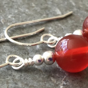 May include: A pair of silver dangle earrings with red carnelian beads. The earrings have a simple design with a small silver bead separating the carnelian bead from the hook.