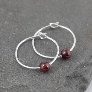 May include: A pair of silver hoop earrings, each featuring a deep red, round gemstone. The earrings are displayed on a dark gray, textured surface, showcasing their simple yet elegant design. The gemstones add a pop of color.