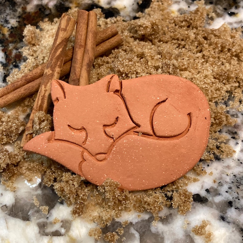 May include: A terracotta-colored ceramic fox figurine curled up in a sleeping position. The figurine is surrounded by brown sugar crystals.