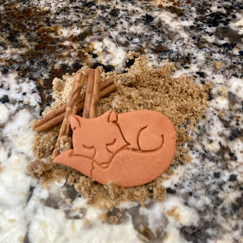 May include: A brown ceramic fox cookie cutter nestled in brown sugar with cinnamon sticks.