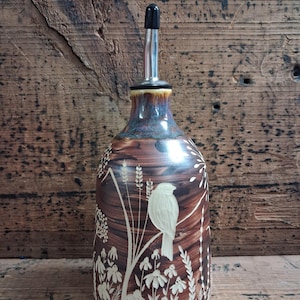 May include: A ceramic oil cruet with a brown glazed finish featuring a white sgraffito design of a bird perched on a stem surrounded by wildflowers and tall grasses. The bottle has a tapered neck and a stainless steel pour spout with a black cap.