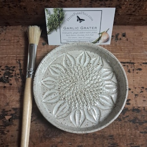 May include: A round, speckled ceramic garlic grater with a sunflower-shaped center, accompanied by a wooden-handled brush. A white product card displays the text HYDEPARKHILL.COM, GARLIC GRATER, and lists uses like garlic, ginger, and citrus zest.